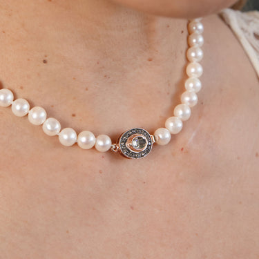 A close-up of a pearl necklace worn on a person's neck, featuring a round rose gold pendant with a central clear gemstone surrounded by small dark stones.